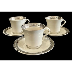 May include: Three white teacups and saucers with silver trim. The cups have a classic shape with a curved handle. The saucers and the rims of the cups are decorated with a silver geometric pattern. The set is displayed on a black background.