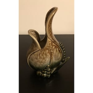 May include: A ceramic vase shaped like a tulip, featuring a curved handle. The vase has a mottled brown and green glaze, with a cluster of small, round details near the base. The vase is on a black surface.