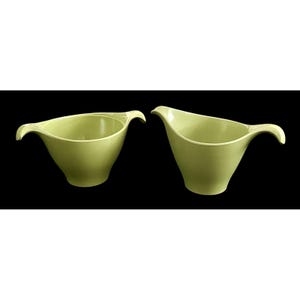 May include: Two vintage, light green serving bowls with a unique, curved handle design. The bowls have a smooth, glossy finish and a classic, mid-century modern aesthetic. The bowls are set against a black background.