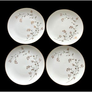 May include: Four white ceramic plates with a gold rim and a floral design in shades of brown and gray. The floral pattern curves across the plate's surface. The plates are arranged on a black background.