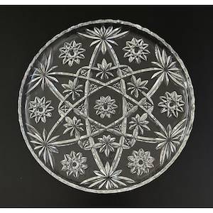 May include: Clear glass serving platter with a starburst and geometric design. The round platter features a detailed pattern of star shapes and geometric lines, perfect for serving appetizers or desserts. The edge has a scalloped design.