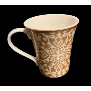 以下が含まれることがあります： 白いハンドルと内側のセラミックコーヒーマグ。 外側は金と白の花柄です。 マグカップはわずかにフレアした形状で、黒い背景に配置されています。