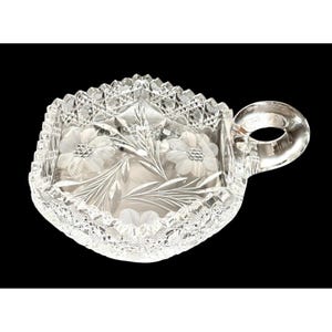 May include: A clear, cut-glass dish with a handle. The dish features a floral design with etched flowers and leaves. The rim has a saw-tooth edge. The handle is a loop shape. The dish is likely vintage.