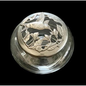 Puede incluir: Un tarro de cristal transparente con una tapa plateada con un diseño de colibrí. La tapa tiene un relieve detallado de un colibrí en vuelo, rodeado de hojas y flores. El tarro es redondo y está sobre un fondo negro.