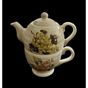Puede incluir: Juego de té individual de cerámica color crema con tetera y taza. El juego está decorado con racimos de uvas verdes y moradas y hojas. La tetera se asienta sobre la taza, creando un diseño compacto y elegante.