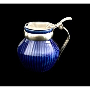 May include: A cobalt blue ceramic creamer with a vertical ribbed texture and a rounded body. It features a polished silver-toned metal rim, a matching hinged lid, and a curved handle extending from the side for easy pouring and handling.