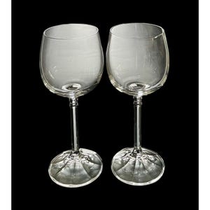 May include: Two clear wine glasses with a rounded bowl and a long stem. The base of each glass has a textured, geometric design. The glasses are set against a black background.