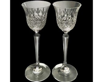May include: Two clear crystal wine glasses with intricate diamond and vertical line patterns. The glasses have long, faceted stems and round bases. The background is solid black, highlighting the clarity and detail of the glassware.