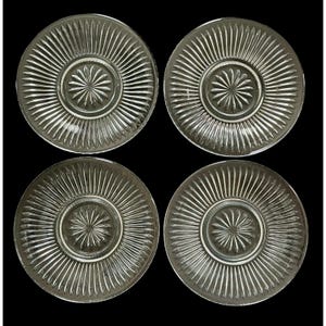 May include: Four clear glass plates with a radial ribbed design and a starburst pattern in the center. The plates are arranged in a square formation against a black background, showcasing their vintage aesthetic.