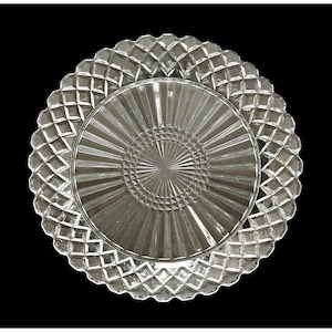 Puede incluir: Plato de servir de cristal transparente con un patrón de diamantes alrededor del borde y un diseño radial en el centro. El plato es circular y se encuentra sobre un fondo negro. Probablemente se utiliza para servir aperitivos o postres.