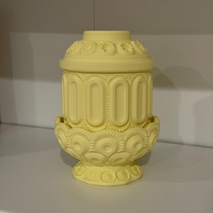 May include: A pale yellow decorative jar with a lid. The jar has a cylindrical body with vertical oval indentations and ornate floral and geometric patterns. The lid and base also feature detailed designs. The jar is made of ceramic or a similar material.