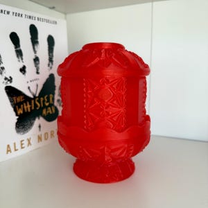 May include: A bright red decorative lantern featuring detailed geometric designs. The lantern is made from a solid material and has a textured surface. The lantern is on a white shelf next to a book titled "The Whisper Man".