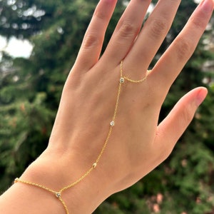 May include: A delicate gold hand chain bracelet with small, sparkling clear stones and a connecting ring. The bracelet is worn on the wrist and connects to a ring worn on the middle finger. The background is a blurred view of green trees.