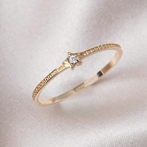 Könnte beinhalten: Ein zarter Goldring mit einem kleinen sternförmigen Diamanten in der Mitte. Die Band ist dünn und hat ein strukturiertes Design.