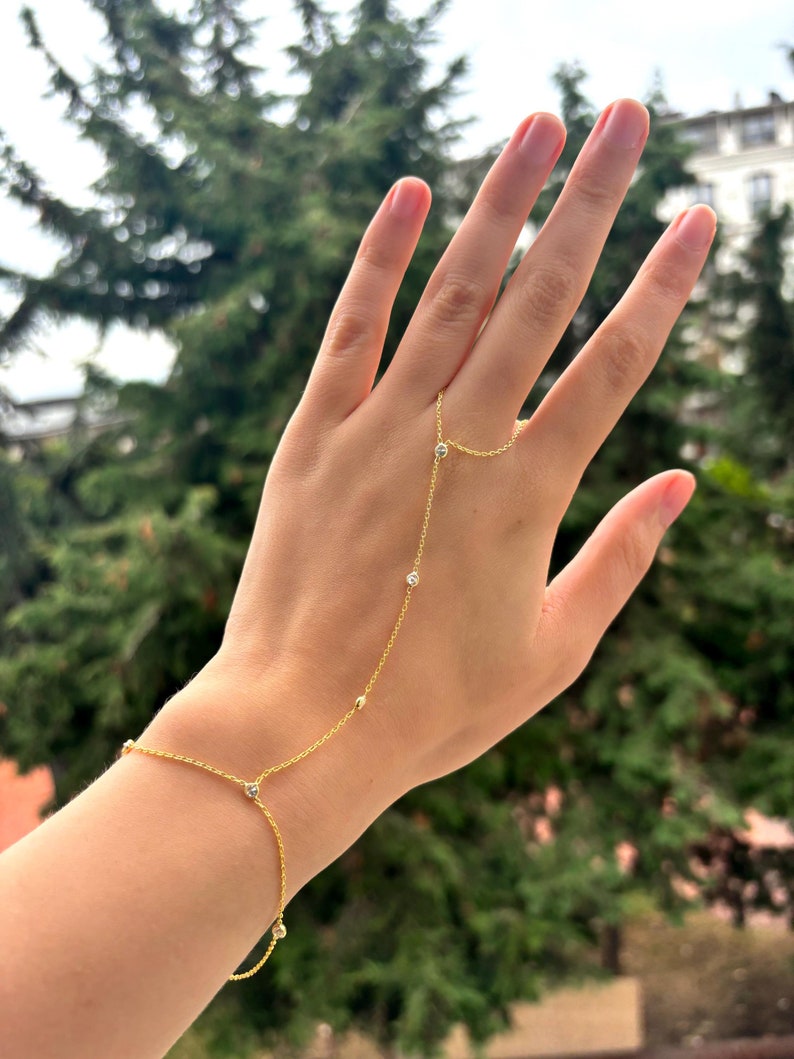 May include: A delicate gold hand chain bracelet with small, sparkling clear stones and a connecting ring. The bracelet is worn on the wrist and connects to a ring worn on the middle finger. The background is a blurred view of green trees.