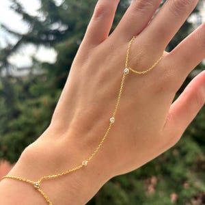May include: A delicate gold hand chain bracelet with small, sparkling gemstones. The bracelet connects from the wrist to the finger, creating a graceful and elegant look. The jewelry is displayed against a blurred green background.