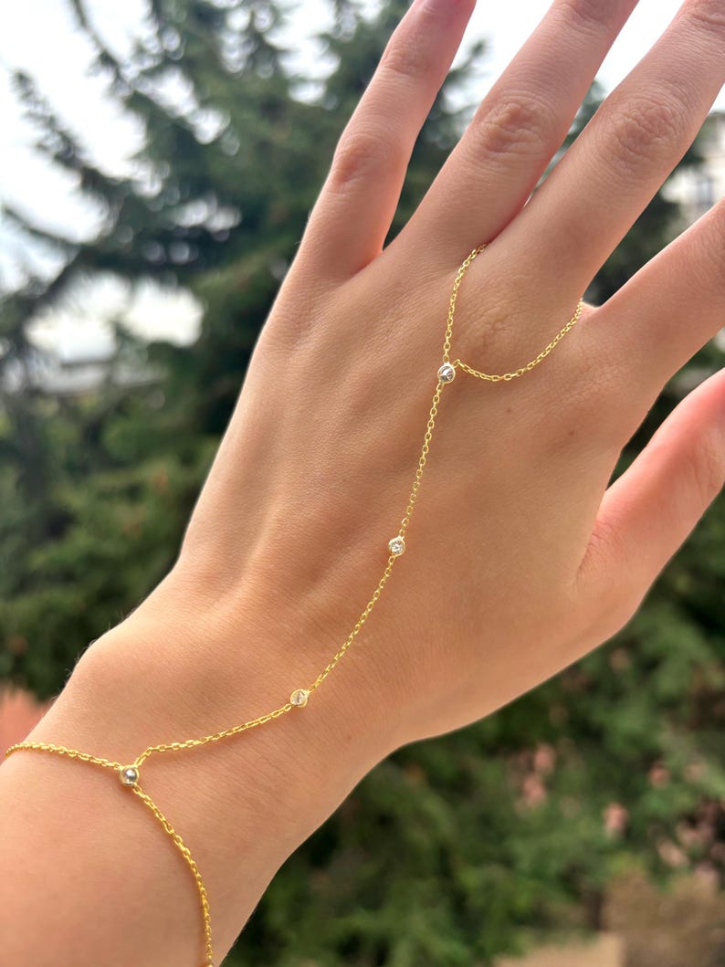 May include: A delicate gold hand chain bracelet with small, sparkling gemstones. The bracelet connects from the wrist to the finger, creating a graceful and elegant look. The jewelry is displayed against a blurred green background.