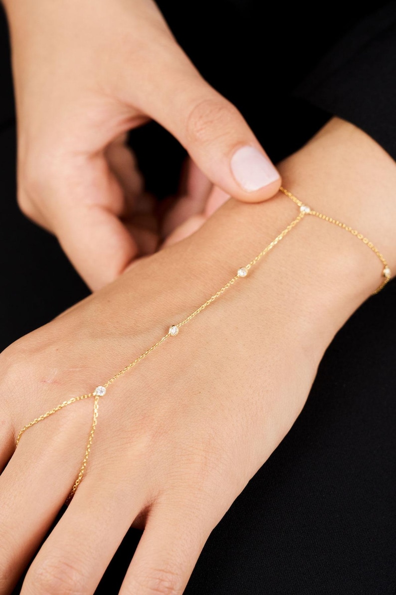 May include: A delicate gold hand chain featuring small, sparkling gemstones. The chain connects to the finger and wrist, creating an elegant and minimalist jewelry piece. The hand chain is shown against a dark background.