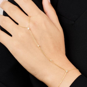 May include: A delicate gold hand chain with small, sparkling gemstones. The chain connects a bracelet to a ring, with a small gem at the center of the hand. The jewelry is displayed against a black background.