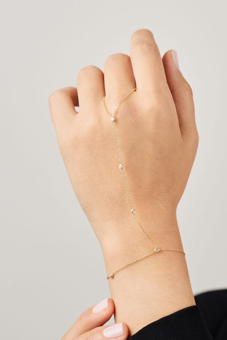 May include: A delicate gold hand chain bracelet featuring small, sparkling gemstones. The bracelet connects from the ring finger to the wrist with a fine chain, creating an elegant and minimalist look. The jewelry is shown on a person's hand.