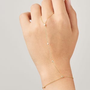 May include: A delicate gold hand chain bracelet featuring small, sparkling gemstones. The bracelet connects from the ring finger to the wrist with a fine chain, creating an elegant and minimalist look. The jewelry is shown on a person's hand.