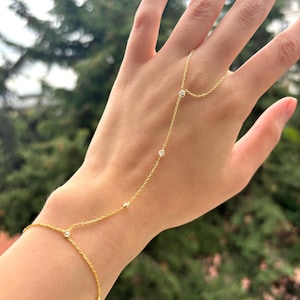 May include: A delicate gold hand chain bracelet with small, clear gemstones. The bracelet connects from the wrist to the middle finger with a fine chain. The jewelry is displayed against a blurred green background.