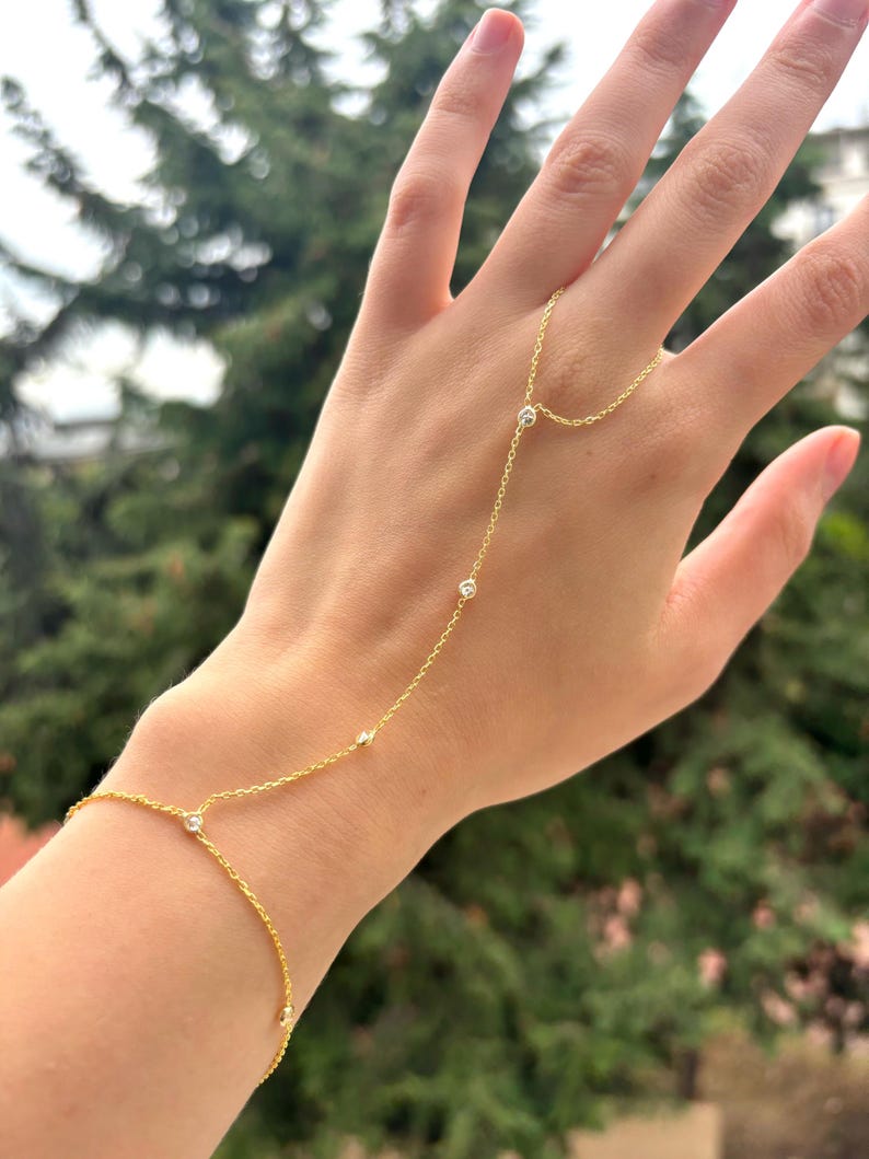 May include: A delicate gold hand chain bracelet with small, clear gemstones. The bracelet connects from the wrist to the middle finger with a fine chain. The jewelry is displayed against a blurred green background.