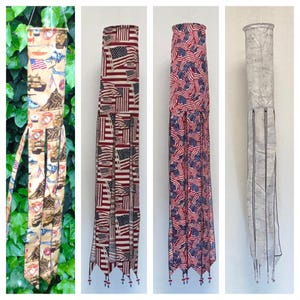May include: Four decorative wind socks. One features military-themed imagery, while the others display American flags in various patterns. The wind socks are long and cylindrical, with decorative tassels at the bottom. They are designed to hang and catch the wind.