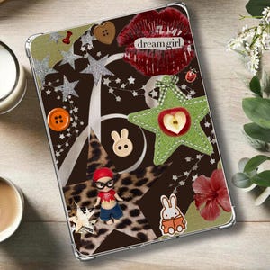 Könnte beinhalten: Eine dekorative Tablet-Hülle mit braunem Hintergrund und verschiedenen Verzierungen. Zu den Merkmalen gehören ein rotes Lippen-Design mit dem Text "dream girl", ein grüner Stern mit einem Apfel und eine kleine Figur. Die Hülle ist von einem transparenten Rand umgeben.