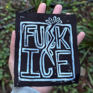 Może przedstawiać: Czarny kwadratowy nadruk z napisem "FUCK ICE" w jasnoniebieskich literach blokowych. Tekst jest obramowany jasnoniebieską obwódką i stylizowanym kwiatowym wzorem. Nadruk jest trzymany w dłoni na rozmytym zielonym tle.