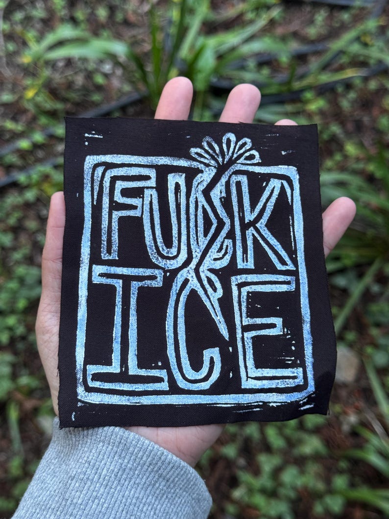 May include: A black square print featuring the words "FUCK ICE" in light blue block letters. The text is framed by a light blue border and a stylized floral design. The print is held in a hand against a blurred green background.