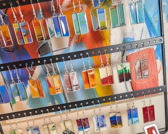 May include: A display of rectangular dangle earrings with silver hooks. The earrings feature colorful abstract designs in shades of blue, orange, red, and green. They are arranged on a black metal earring stand against a colorful painted background.