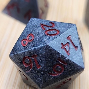 May include: Close-up of a black and gray D20 die with red numbering. The twenty-sided die is textured and has the numbers 8, 16, 19, 14, and 20 visible. The die is likely used for tabletop role-playing games.