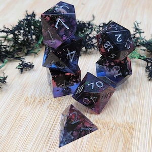 Pode incluir: Um conjunto de sete dados poliédricos com uma base de resina escura e translúcida, com inclusões em tons de roxo, azul e preto. Cada dado possui arestas afiadas e números gravados na cor prata para garantir uma visibilidade clara ao jogar.
