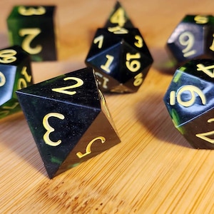 Könnte beinhalten: Eine Sammlung von Würfeln in verschiedenen Formen mit einer schwarz-grünen Marmorierung und gelben Zahlen. Die Würfel liegen auf einer Holzoberfläche und zeigen ihre unterschiedlichen Seiten und Zahlen.