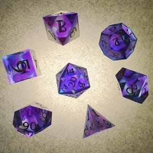 Puede incluir: Conjunto de siete dados iridiscentes de color morado y azul con números negros. Los dados son un d4, d6, d8, d10, d%, d12 y d20.