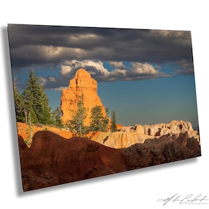 May include: A landscape photograph featuring a towering orange rock formation under a dramatic sky. The scene includes green trees, rocky terrain, and a mix of dark and light clouds, creating a contrast of colors and textures. The image is a print.