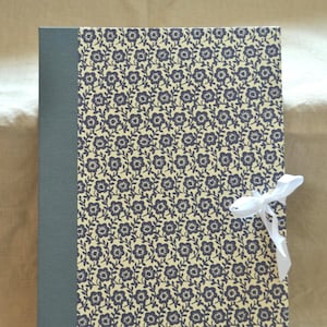 Könnte beinhalten: Eine rechteckige Dokumentenmappe mit einem dunkelgrauen Buchrücken und einem cremefarbenen Einband mit einem sich wiederholenden dunkelblauen Blumenmuster. Die Mappe wird an der rechten Seite mit einer weißen Schleife aus dünnem Stoff verschlossen.