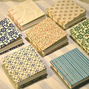 Könnte beinhalten: Sieben quadratische Notizbücher mit sichtbarer Fadenheftung und verschiedenen gemusterten Stoffeinbänden, darunter blaue Blumenmuster, beige Botanikmotive, goldene Geometrie, rosa Blumen, grüne Blumen, blaue Geometrie und blaue Streifen.