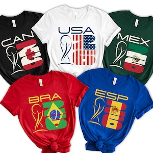 May include: Five short-sleeved t-shirts in black, white, red, blue, and green. Each shirt features a country's flag design with the country's abbreviation and the number 2026. The shirts are for Canada, USA, Brazil, Spain, and Mexico.