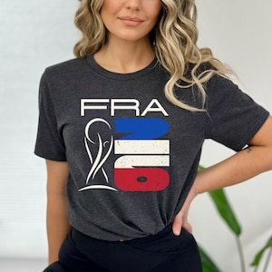 May include: Dark gray t-shirt with the word "FRA" in white block letters above a graphic in the colors of the French flag: blue, white, and red. The graphic includes a stylized image of a person. The shirt is short-sleeved.