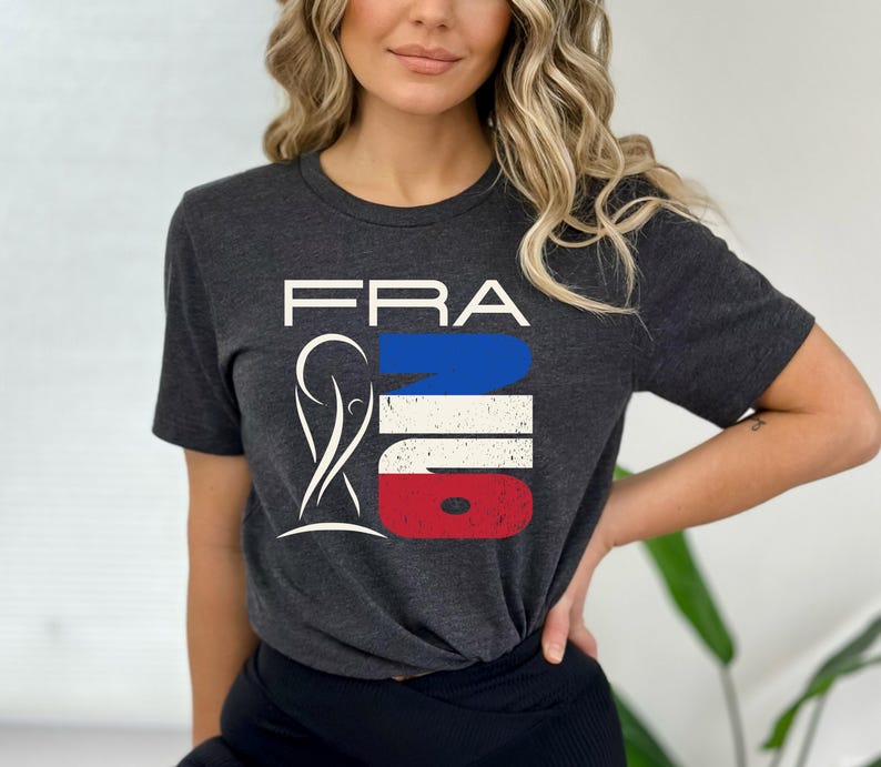 May include: Dark gray t-shirt with the word "FRA" in white block letters above a graphic in the colors of the French flag: blue, white, and red. The graphic includes a stylized image of a person. The shirt is short-sleeved.