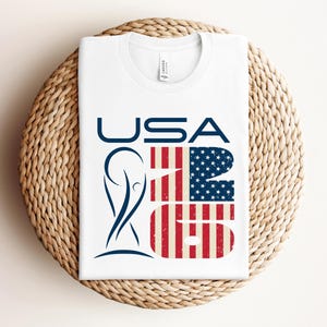 May include: White t-shirt with the word "USA" in navy blue, and the numbers "26" in a design that resembles the American flag. A navy blue graphic is on the left side of the shirt. The shirt is folded on a woven, circular surface.