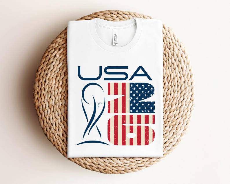 May include: White t-shirt with the word "USA" in navy blue, and the numbers "26" in a design that resembles the American flag. A navy blue graphic is on the left side of the shirt. The shirt is folded on a woven, circular surface.