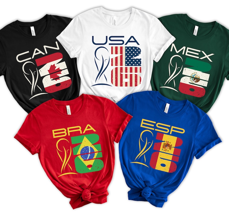 May include: Five short-sleeved t-shirts in black, white, red, blue, and green. Each shirt features a country's flag design with the country's abbreviation and the number 2026. The shirts are for Canada, USA, Brazil, Spain, and Mexico.