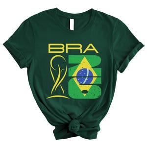 May include: Dark green t-shirt with the word "BRA" in yellow block letters above a stylized yellow figure and a green and yellow design with a blue and white circle. The shirt is knotted at the bottom.