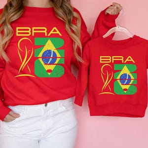 May include: Red sweatshirts with the word "BRA" in yellow block letters and a graphic of the Brazilian flag. One sweatshirt is adult-sized, and the other is child-sized. The sweatshirts are displayed against a pink background.