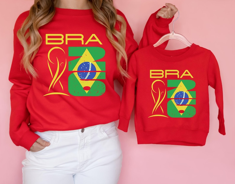 May include: Red sweatshirts with the word "BRA" in yellow block letters and a graphic of the Brazilian flag. One sweatshirt is adult-sized, and the other is child-sized. The sweatshirts are displayed against a pink background.