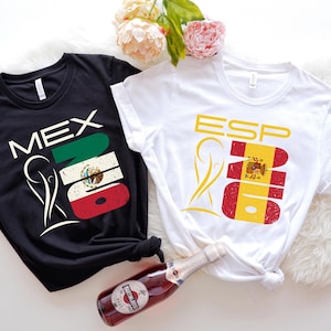 May include: Two t-shirts, one black and one white, with flag-themed designs. The black shirt says "MEX" with the Mexican flag, and the white shirt says "ESP" with the Spanish flag. A bottle of sparkling wine and flowers are in the image.