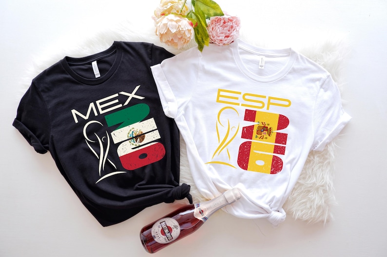 May include: Two t-shirts, one black and one white, with flag-themed designs. The black shirt says "MEX" with the Mexican flag, and the white shirt says "ESP" with the Spanish flag. A bottle of sparkling wine and flowers are in the image.